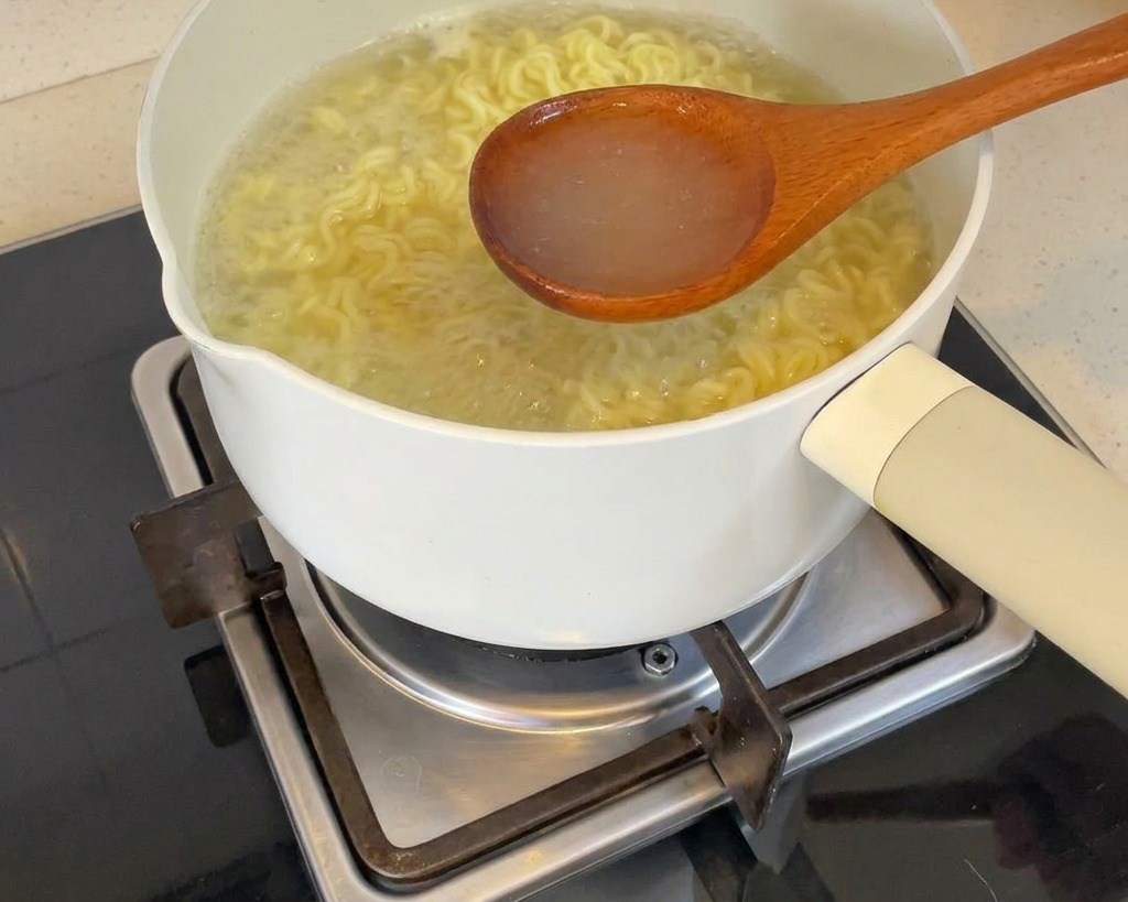 Un mestolo di legno che raccoglie lacqua di cottura dei noodle bollente da una pentola bianca.