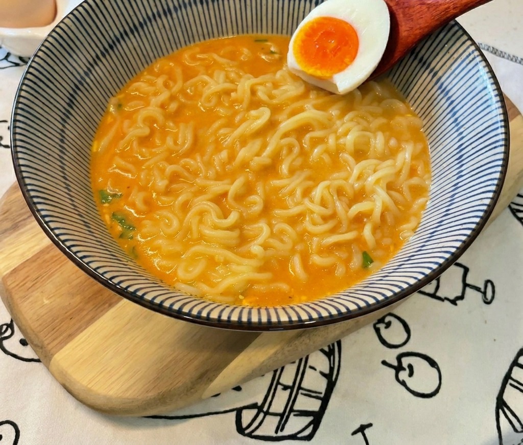 Un cucchiaio di legno adagia mezzo uovo sodo con il tuorlo arancione brillante sopra i noodle.