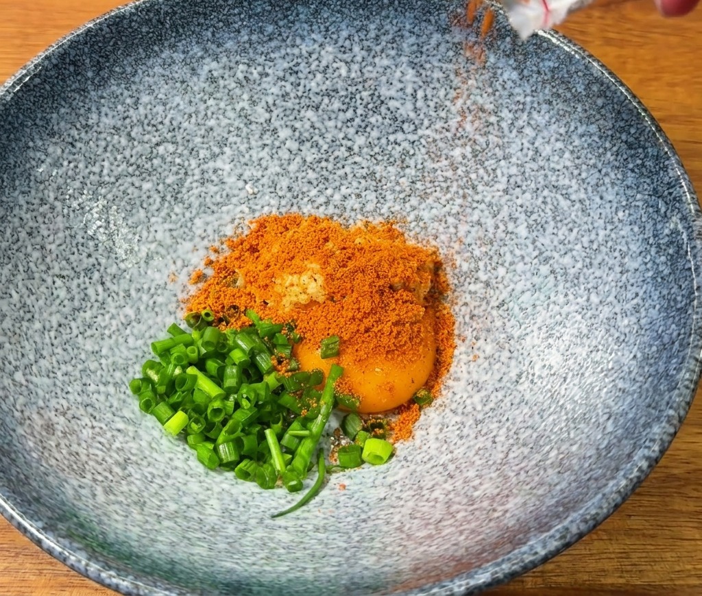 Condimento rosso per ramen versato in una ciotola blu con un tuorlo duovo e cipollotti freschi.