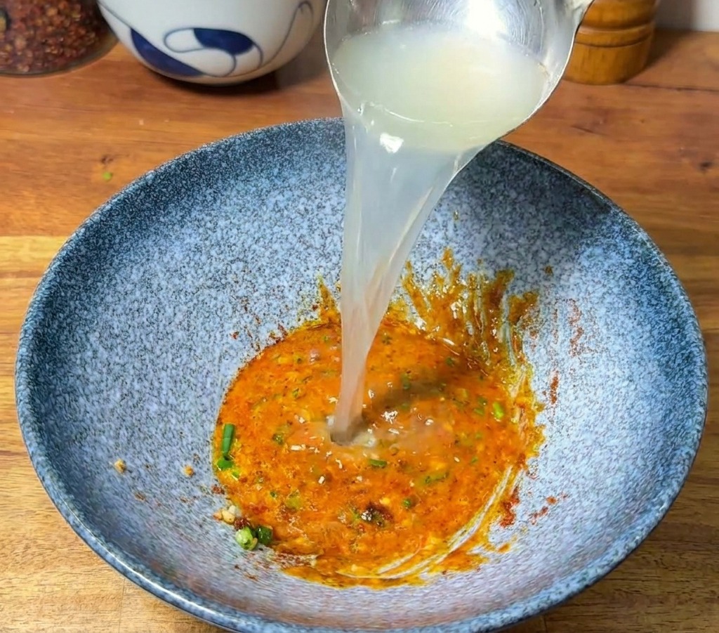 Un mestolo dargento versa acqua bollente fumante in una ciotola sopra la pasta aromatica.