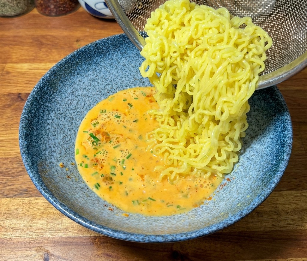 Noodles gialli cotti che vengono trasferiti da un colino a una ciotola di brodo arancione cremoso.
