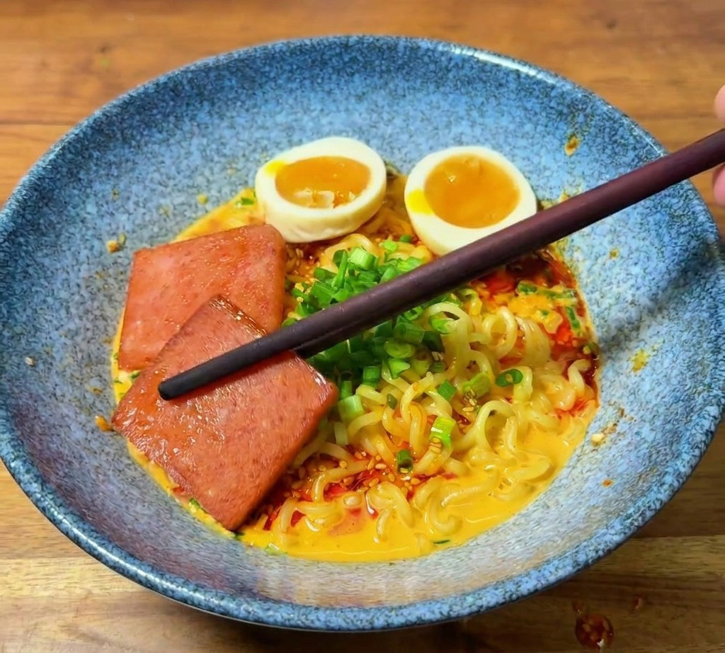 Bacchette che posizionano una fetta di carne rosolata in una ciotola di ramen già guarnita.