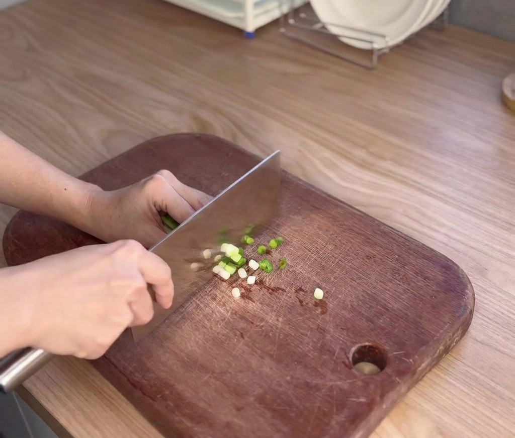 Una persona utilizza un grosso coltello da cucina per affettare finemente dei cipollotti freschi su un tagliere di legno scuro.
