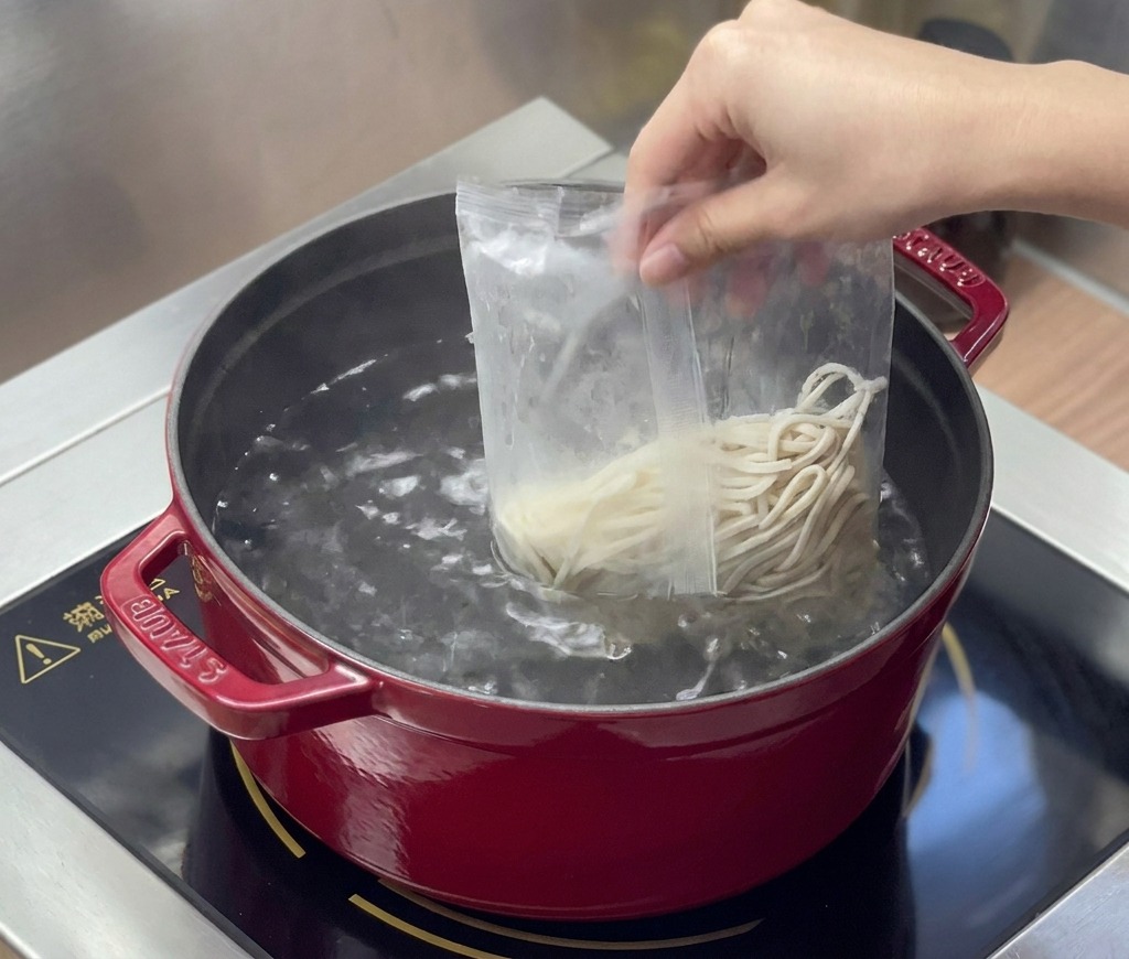Noodle freschi per ramen vengono versati da un sacchetto di plastica trasparente in una pentola rossa contenente acqua in ebollizione.
