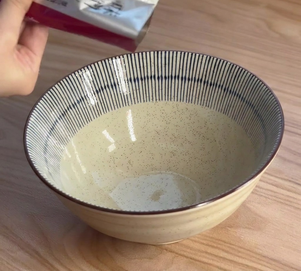 Una mano spreme il concentrato per brodo al miso da una bustina argentata in una ciotola di ceramica a strisce bianche e blu.