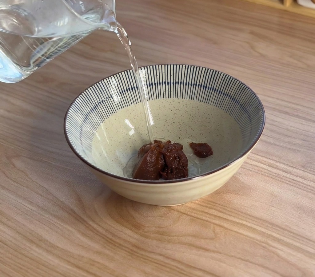 Il vapore sale mentre lacqua bollente viene versata da un misurino in una ciotola di ceramica con dentro la pasta di miso scura.