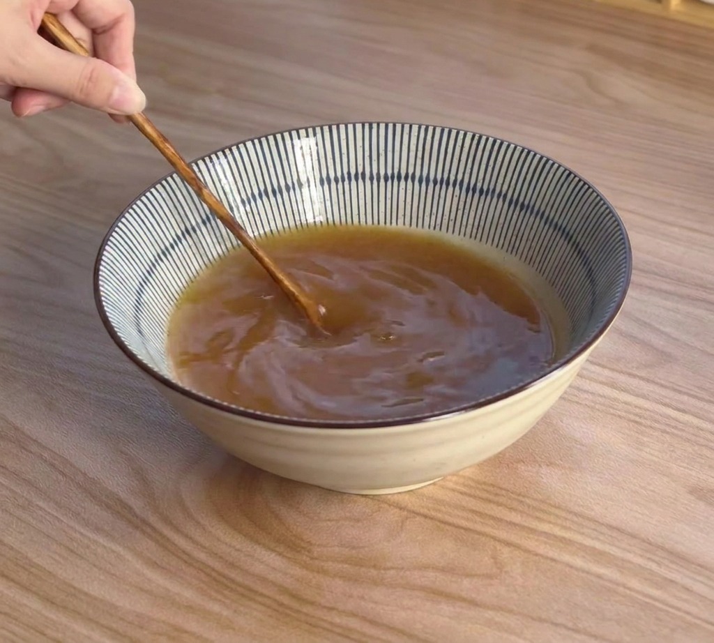 Una mano usa un cucchiaio di legno dal manico lungo per mescolare un ricco brodo marroncino al miso in una ciotola a strisce.