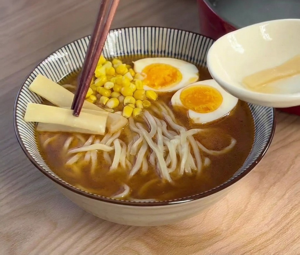 Bacchette in legno scuro posano sottili fette chiare di germogli di bambù in una ciotola di ramen con uovo e mais.