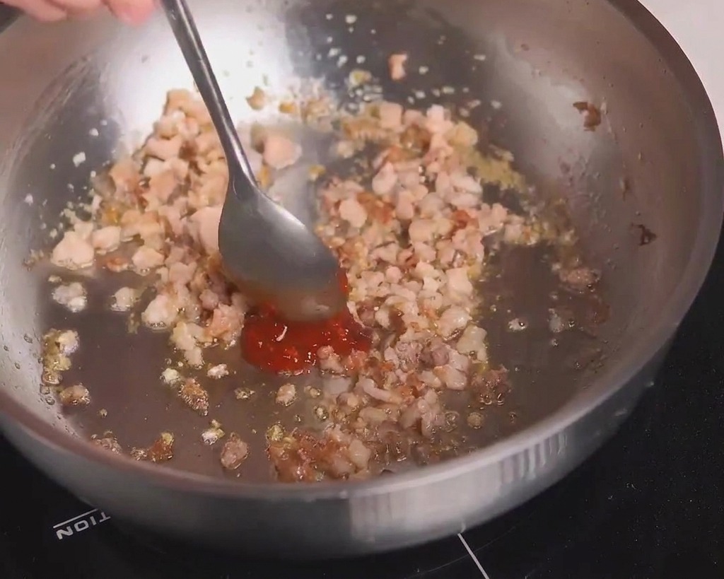 Un cucchiaio di metallo aggiunge una dose di pasta di fagioli piccante rossa alla carne ben rosolata.