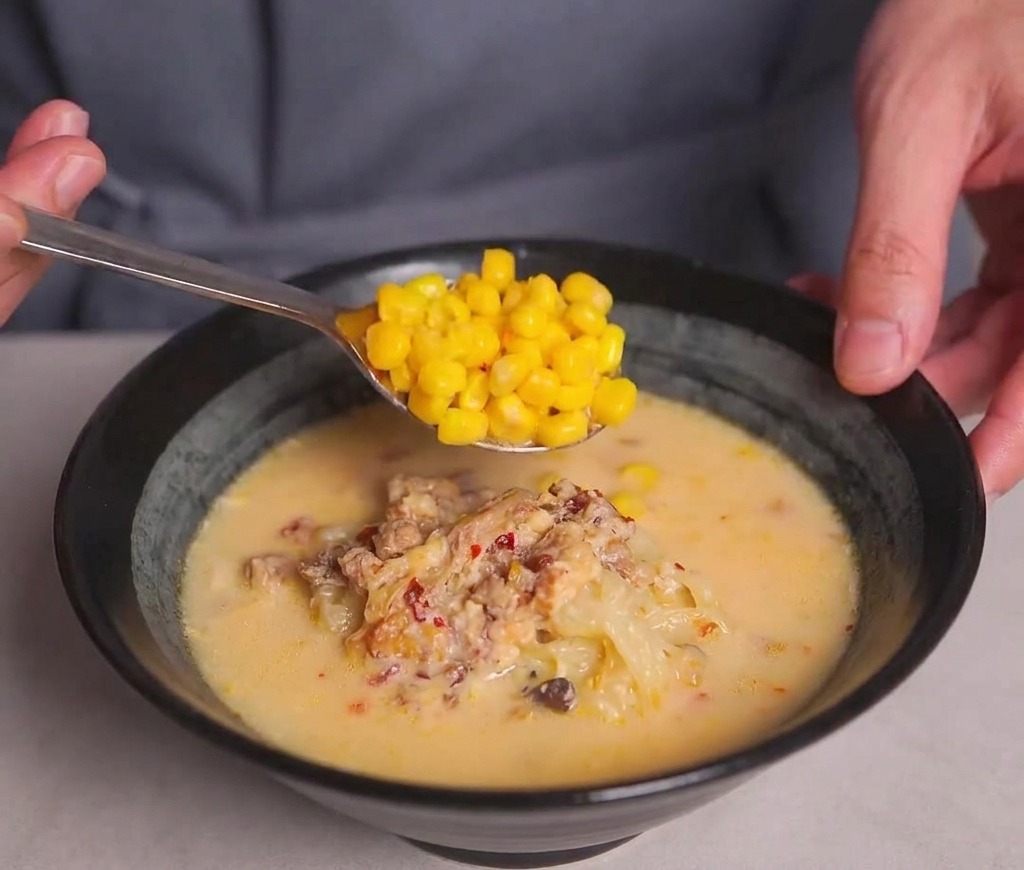 Un cucchiaio di metallo che adagia chicchi di mais dorati sulla superficie di una ciotola di ramen.