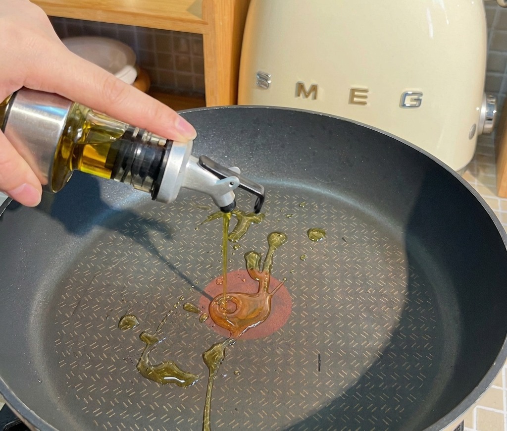 Olio di semi versato da un dosatore in una padella antiaderente nera sul piano cottura.