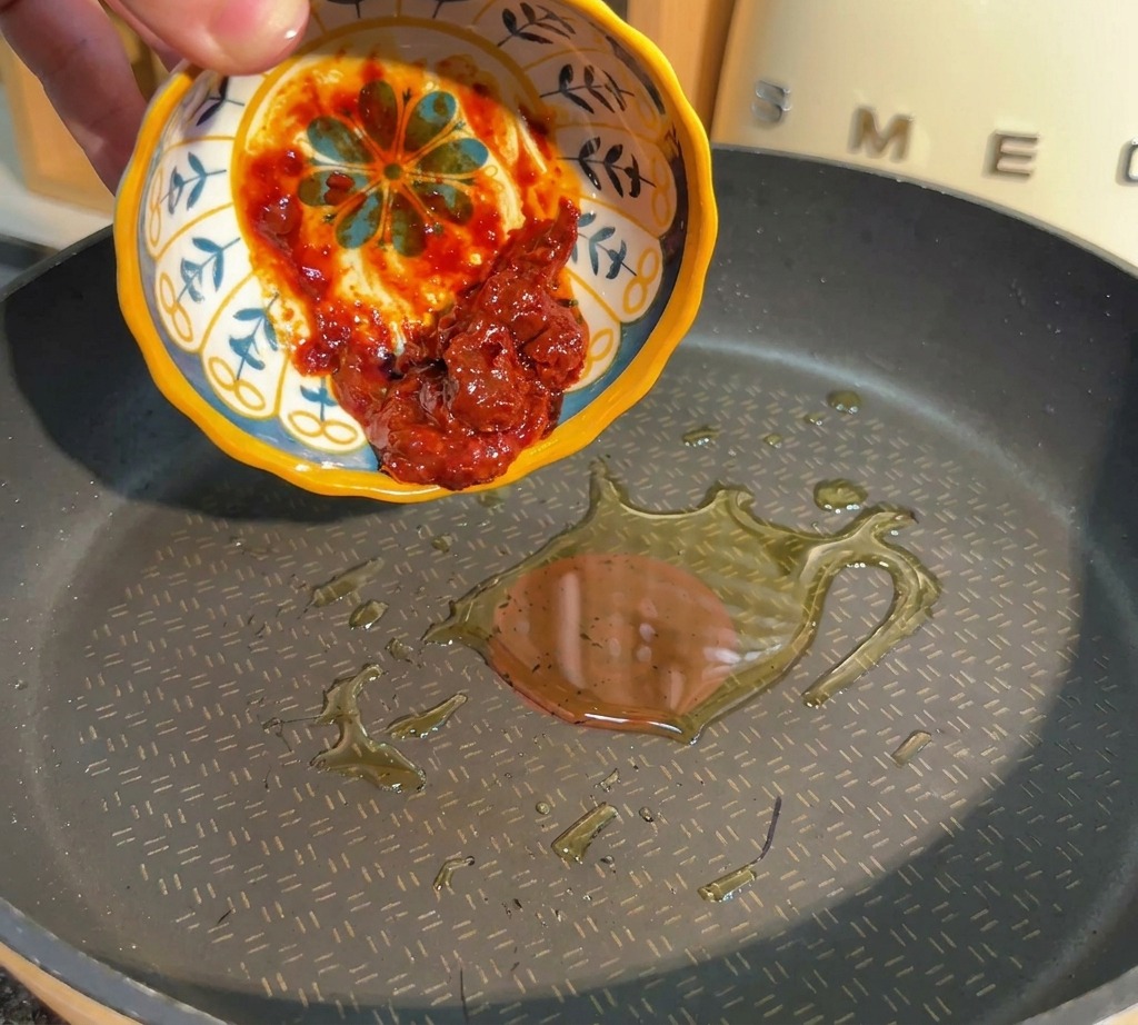 Una mano versa della pasta di fagioli rossa piccante da una ciotolina in una padella con olio caldo.