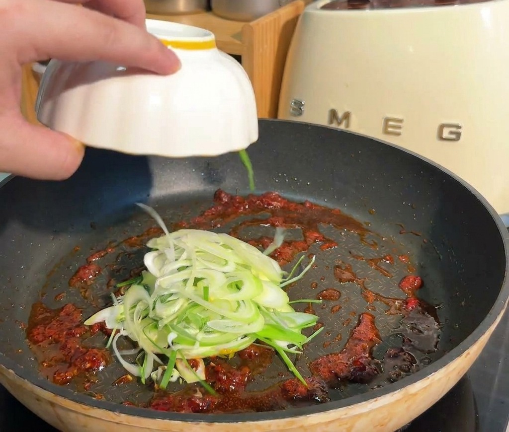 Cipollotti affettati aggiunti da una ciotola bianca a una padella con base di salsa rossa piccante.