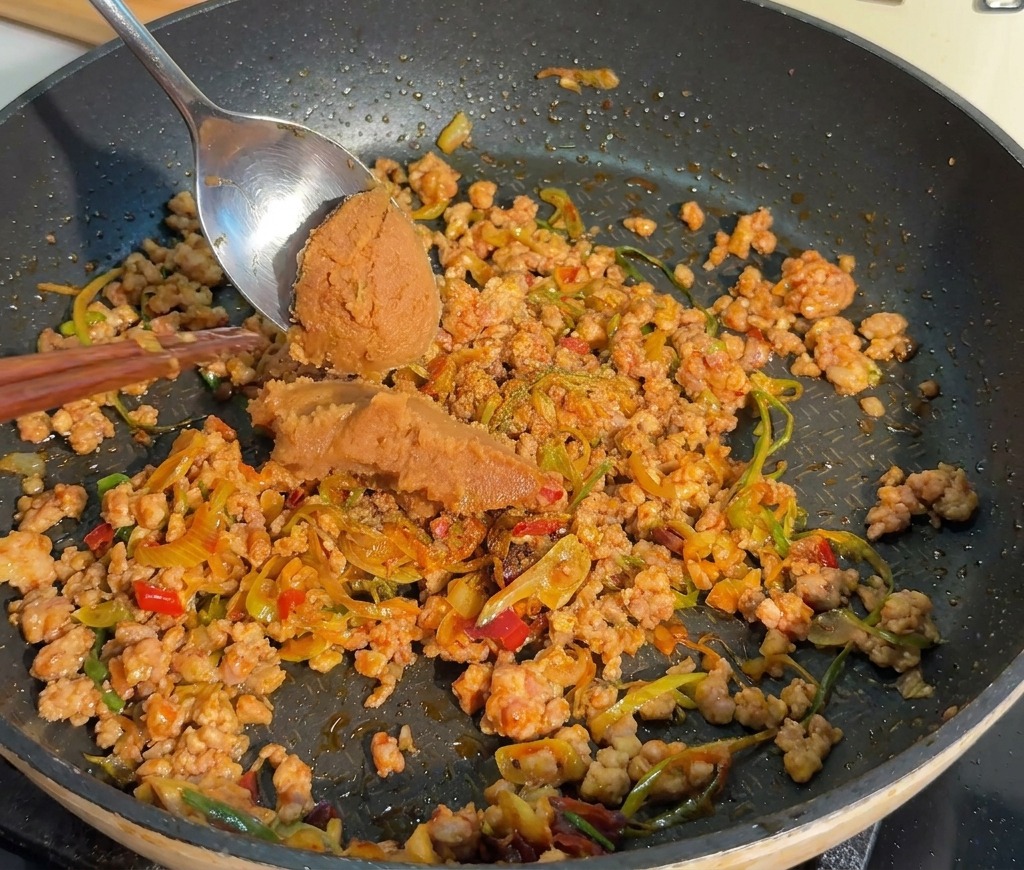 Un cucchiaio di metallo aggiunge una porzione di denso miso rosso alla carne macinata cotta.