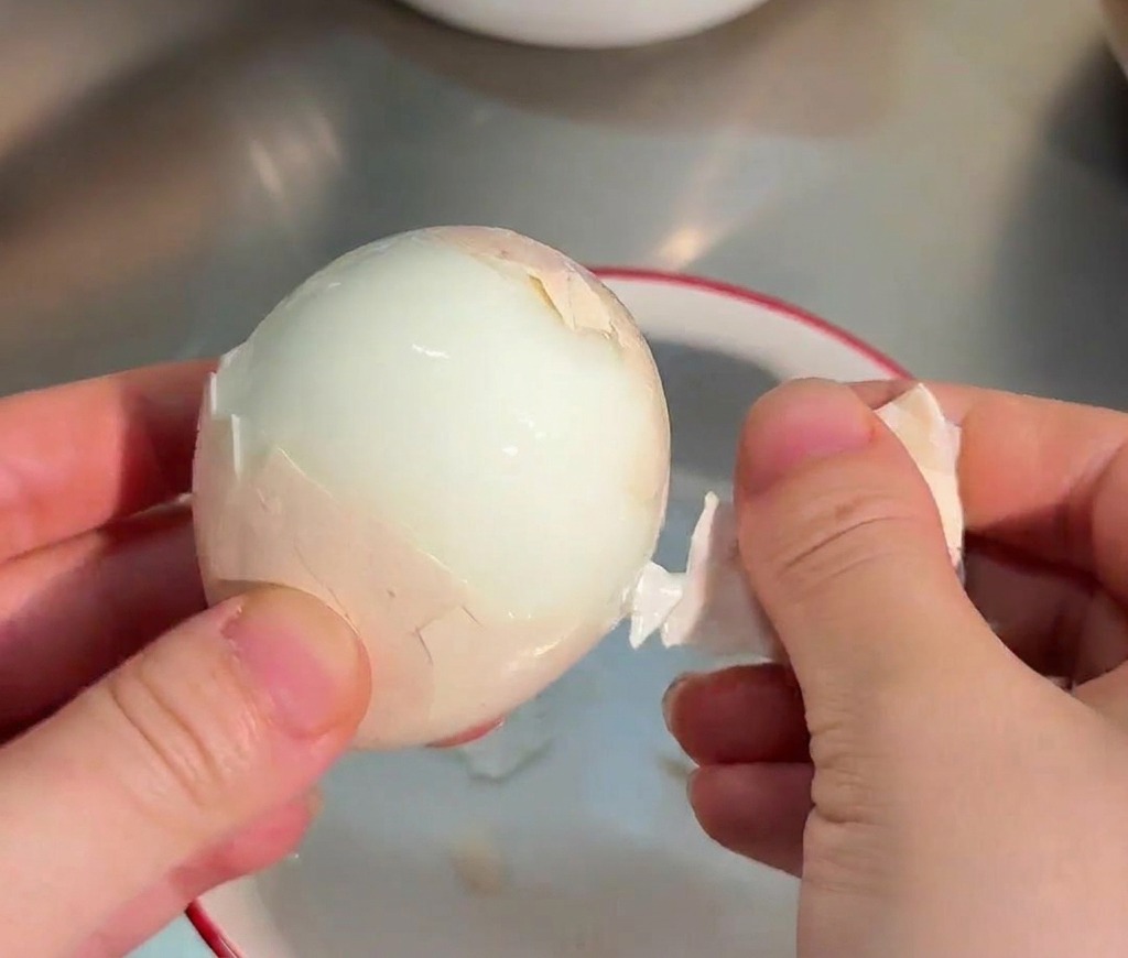 Mani che sbucciano con cura un uovo sodo liscio sopra una ciotola dacqua.