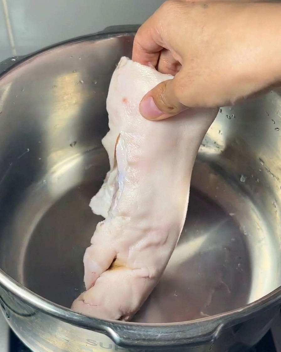 Una mano posiziona un piedino di maiale crudo in una pentola dacciaio insieme a un osso midollare e zampe di gallina.