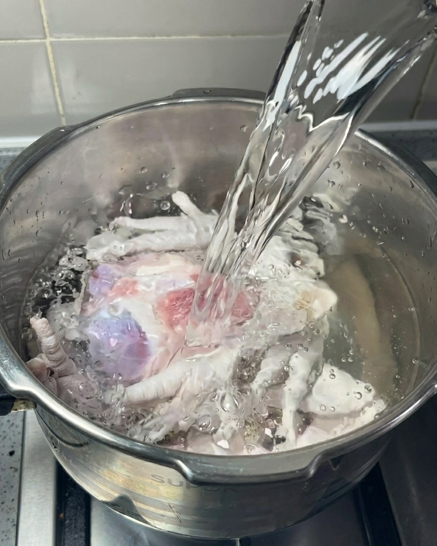 Un getto dacqua limpida viene versato in una pentola dacciaio piena di ossa di maiale crude e zampe di gallina.