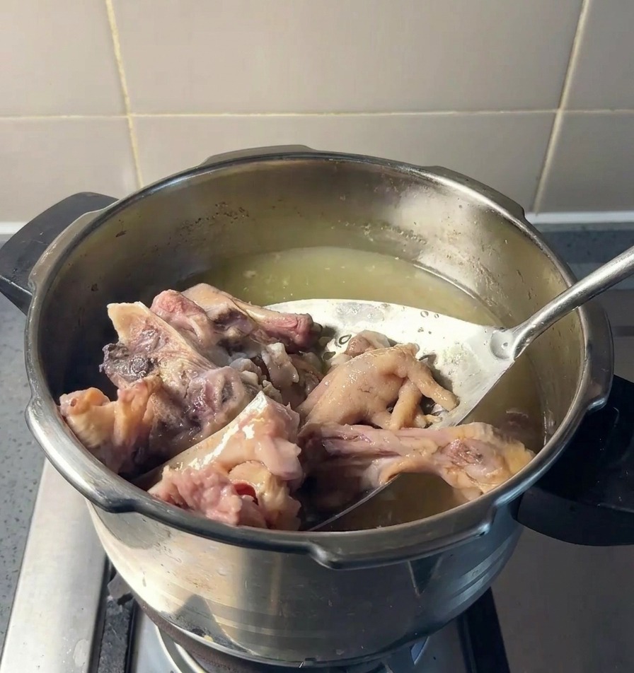 Una schiumarola solleva grandi pezzi di ossa di maiale cotte e zampe di gallina da una pentola di brodo giallastro.