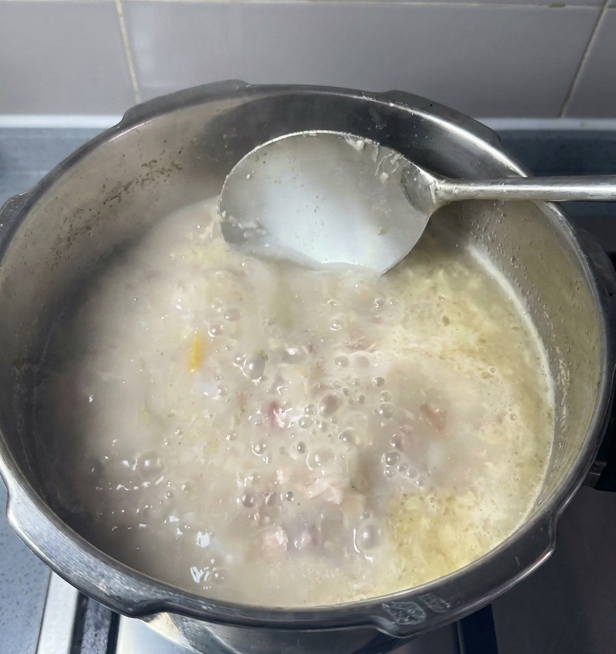 Un mestolo mescola una pentola di brodo di ossa di maiale opaco e bianco latte con pezzetti di carne e grasso.