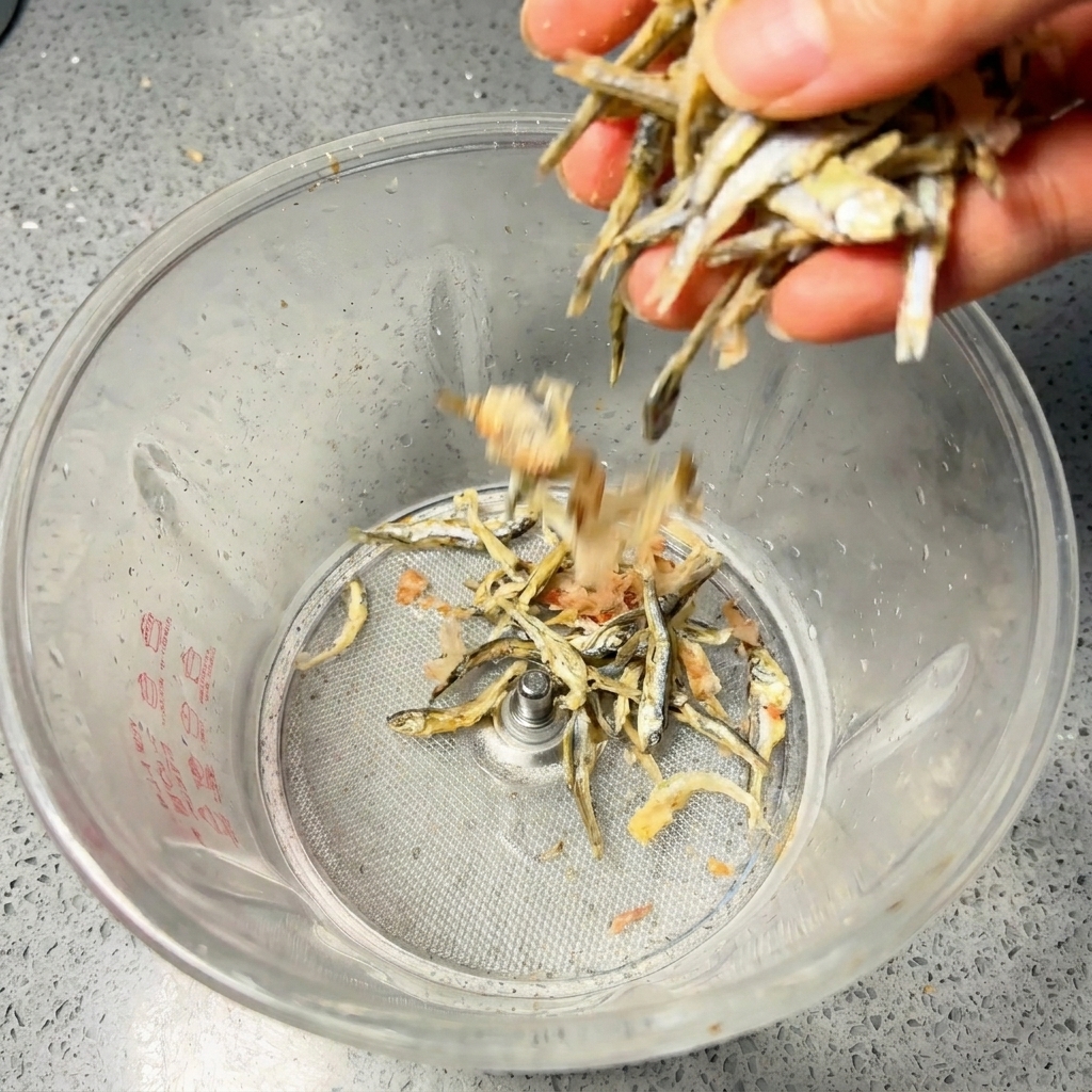 Una mano che versa acciughe essiccate e fiocchi di bonito in un frullatore da cucina trasparente.