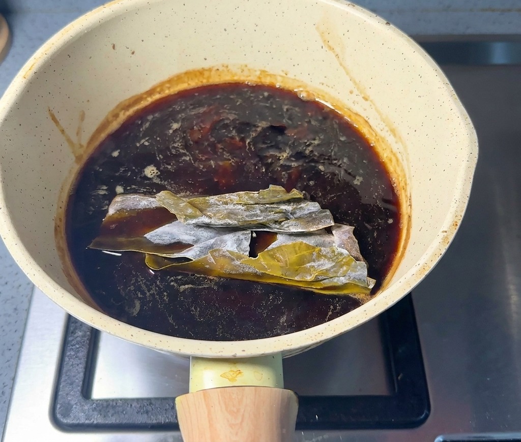 Pezzi di alga kombu essiccata immersi nella salsa scura allinterno di un pentolino chiaro.