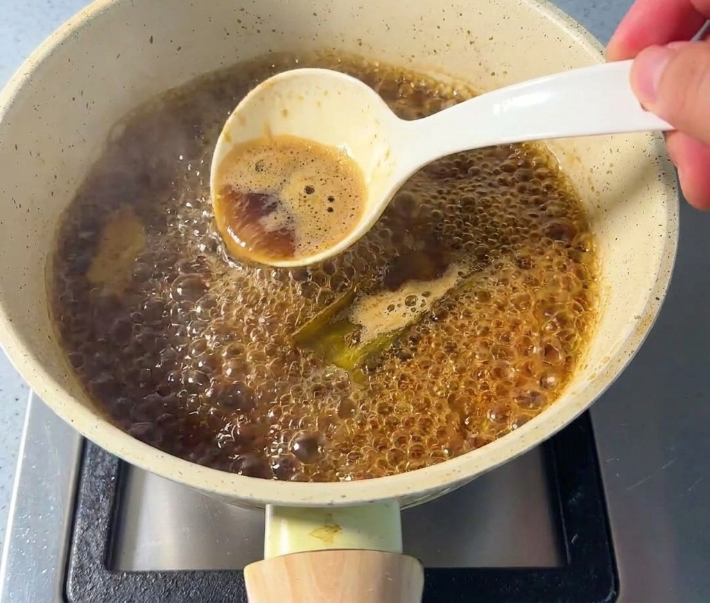 Una mano con un cucchiaio bianco che schiuma la superficie di una salsa scura in ebollizione nel pentolino.