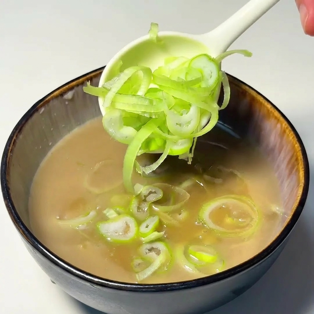 Un cucchiaio bianco che deposita anelli di cipollotto tagliati finemente nel brodo tsukemen.