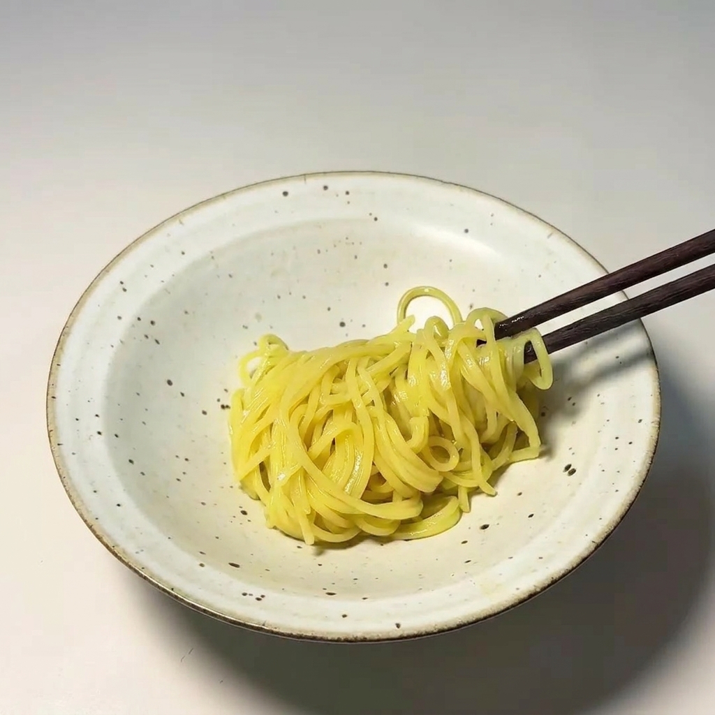 Bacchette di legno scuro che adagiano un mazzetto di noodle gialli in una ciotola di ceramica bianca.