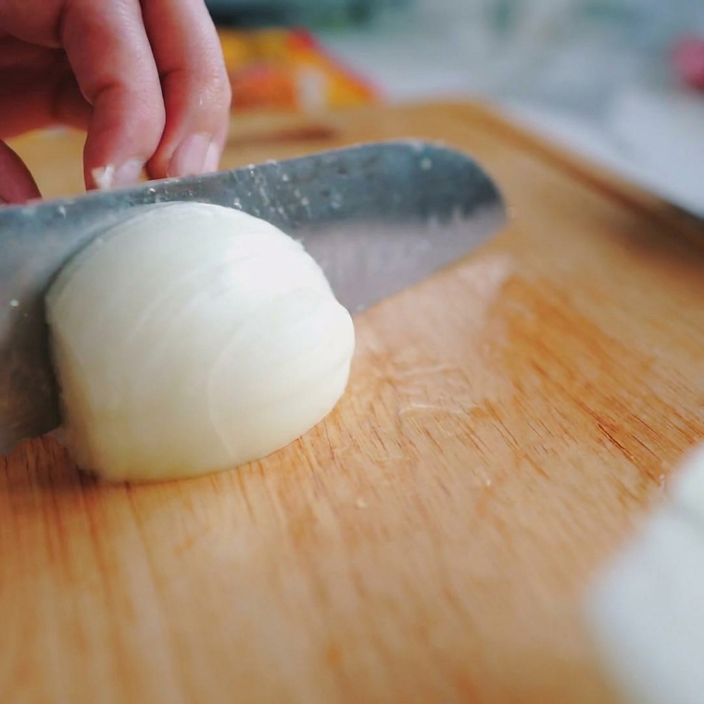 Dettaglio di una mano che affetta una cipolla bianca in strisce sottili e verticali su un tagliere di legno.