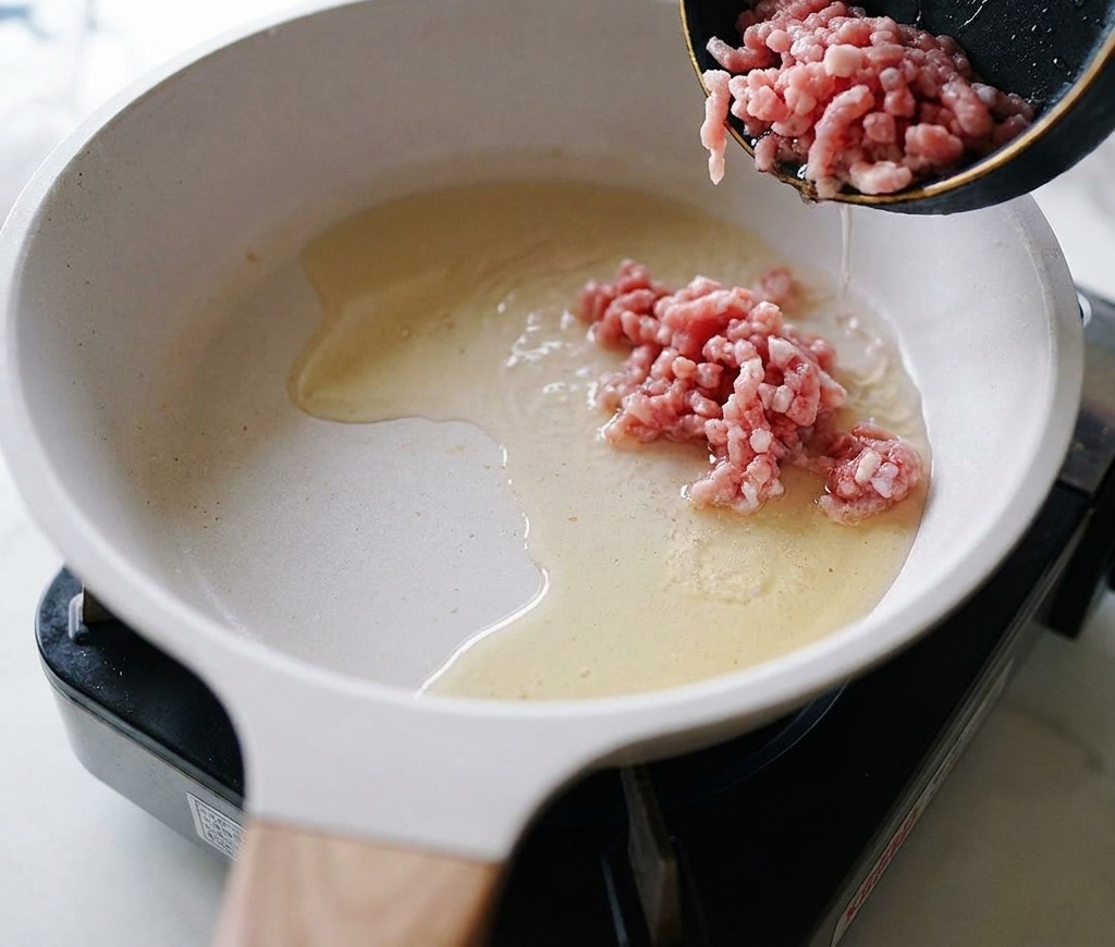 Carne di maiale macinata cruda versata da una padellina in una padella di ceramica con olio caldo.