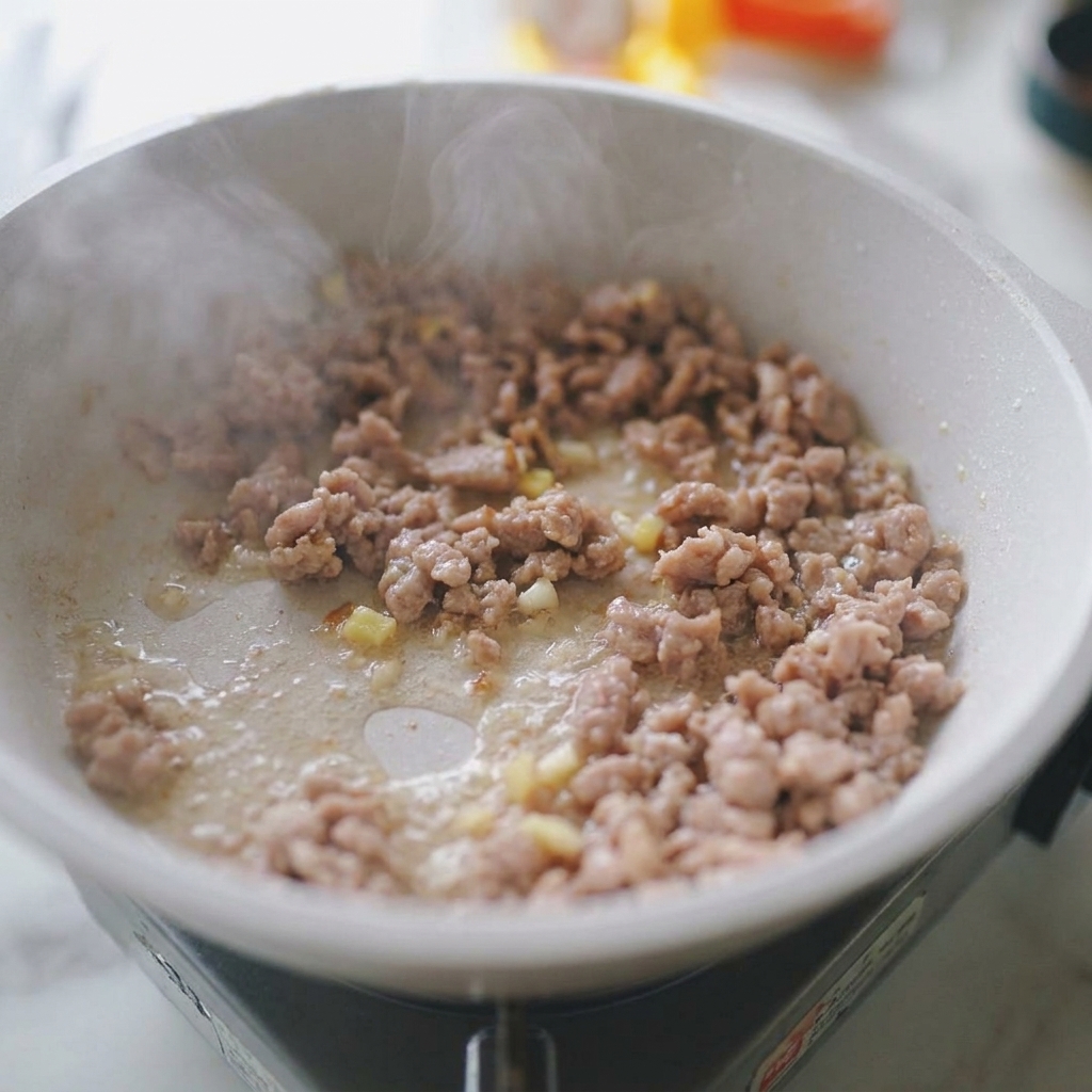 Carne di maiale macinata che salta in padella con pezzetti visibili di aglio e zenzero tritati e vapore che sale.