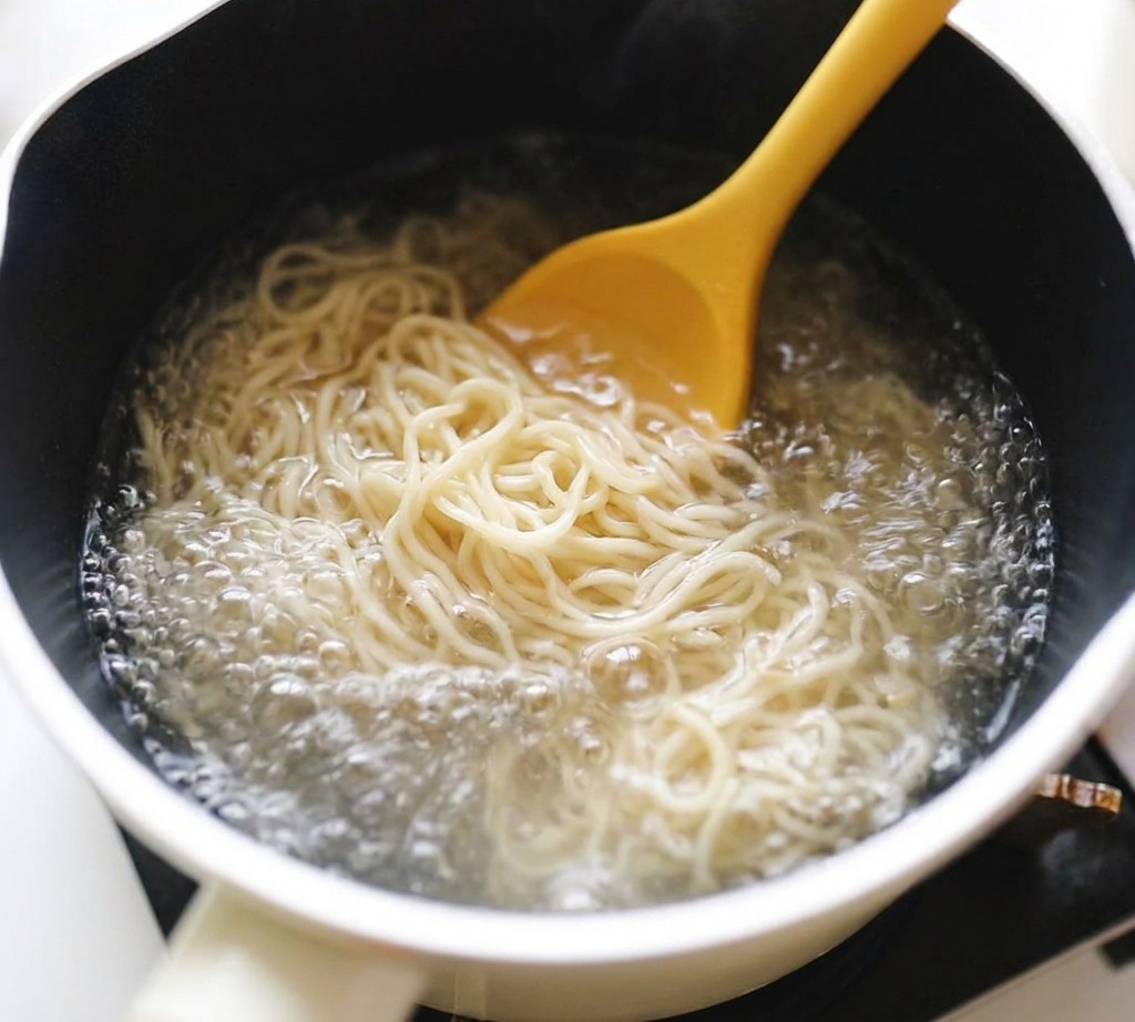Noodles ramen ondulati mescolati con un cucchiaio giallo in una pentola di acqua bollente.