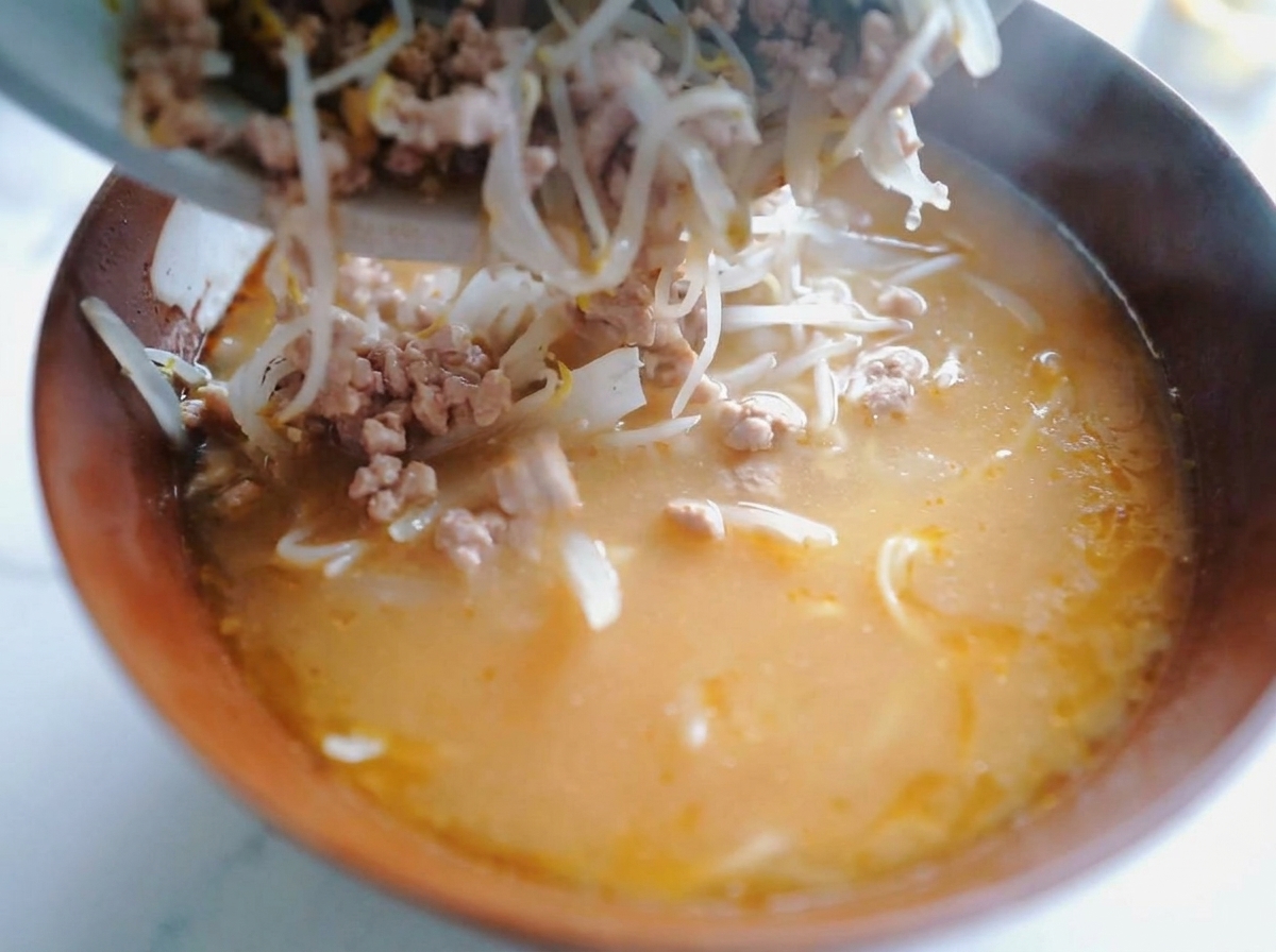 Dettaglio di un cucchiaio che trasferisce il maiale saltato e i germogli di soia sopra i noodles nel brodo di miso.