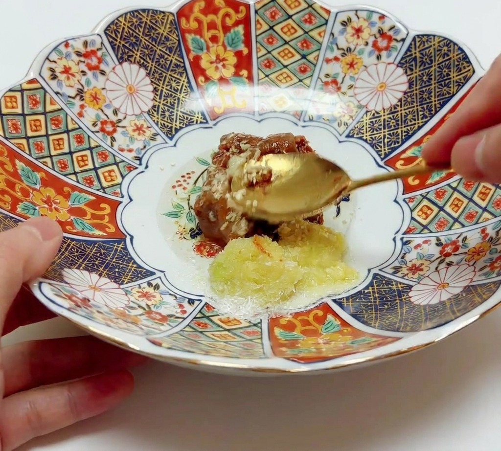 Un cucchiaio dorato che mescola pasta di miso, aromi e olio in una ciotola di ceramica decorata.