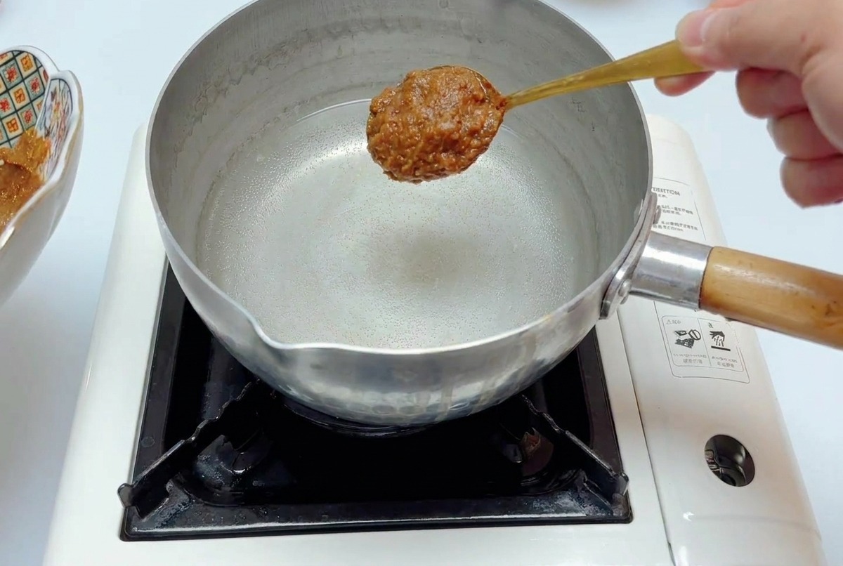 Un cucchiaio di densa salsa al miso sopra una pentola dacciaio contenente acqua limpida su un fornello portatile.