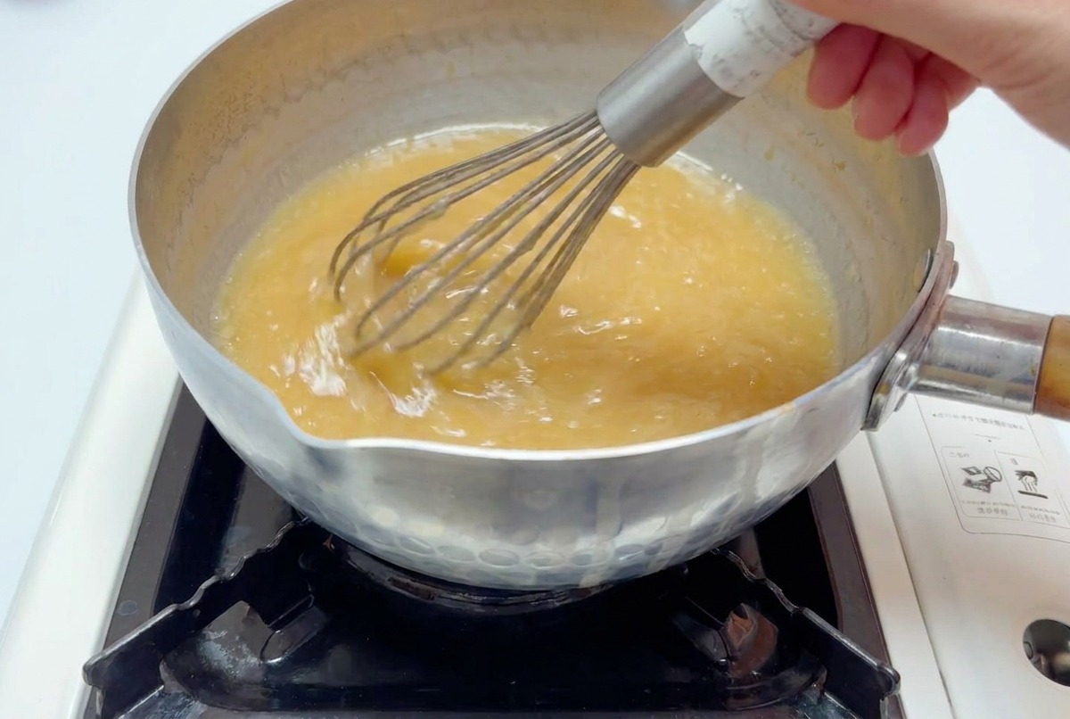 Una frusta di metallo che mescola un brodo marrone chiaro in una pentola sul fuoco fino a completa emulsione.