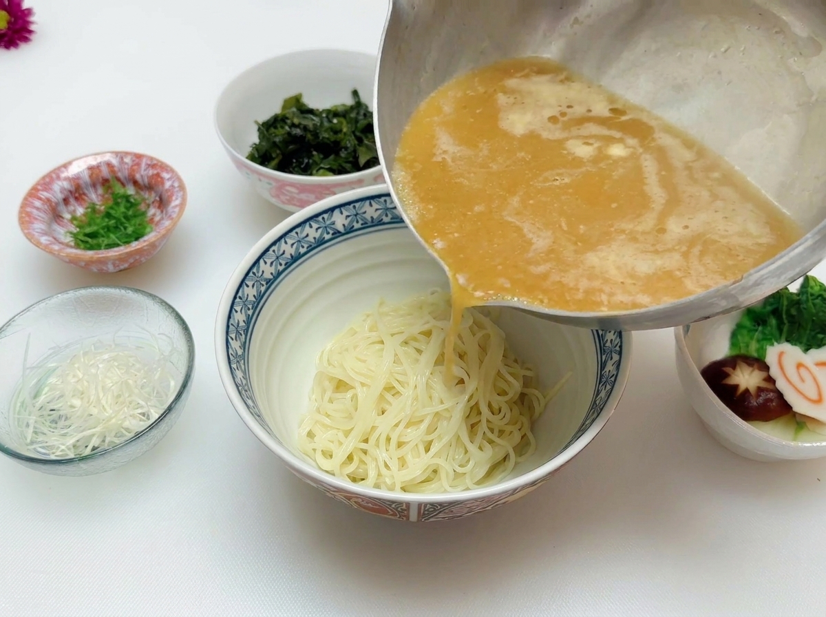 Brodo al miso bollente versato da una pentola dacciaio sopra i noodles già pronti in una ciotola di ceramica.
