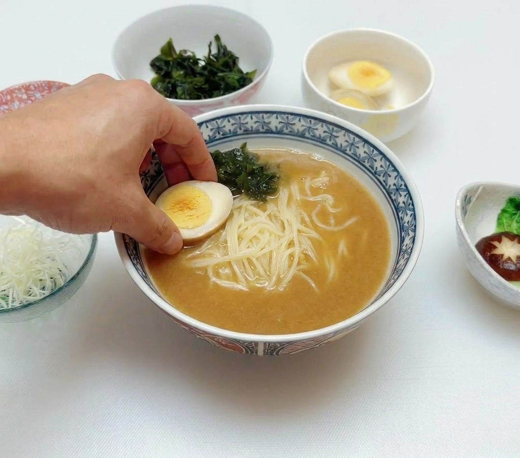 Una mano che posiziona mezzo uovo barzotto in una ciotola di ramen con brodo, noodles e alga wakame.