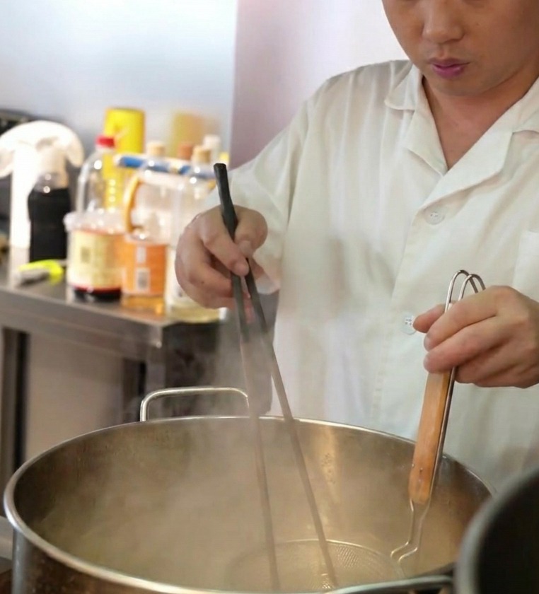 Uno chef che immerge un colino a rete pieno di noodle freschi in una grande pentola di acqua bollente.