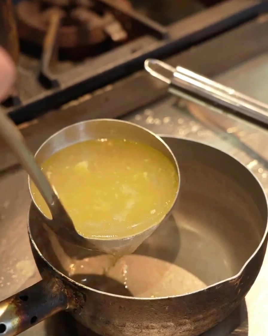 Brodo giallo limpido versato da un mestolo in un piccolo pentolino di metallo sopra un piano da cucina.