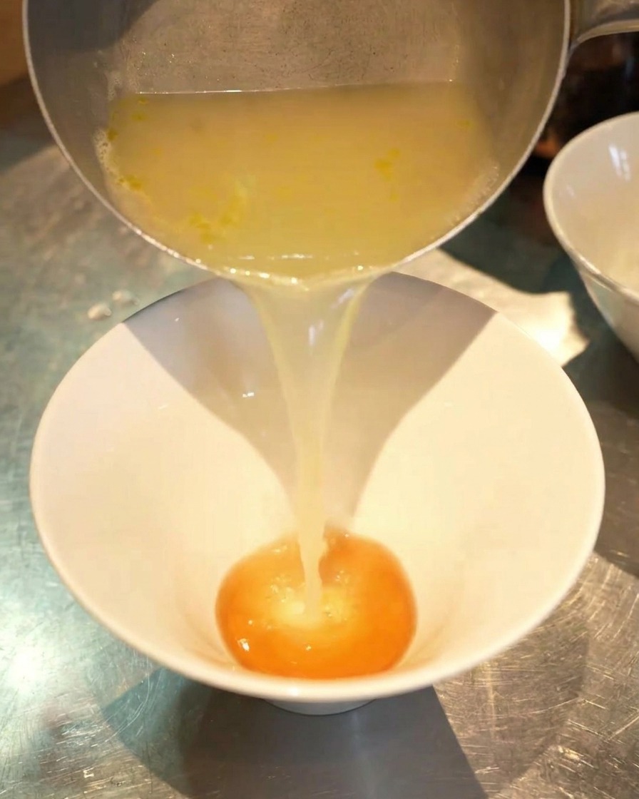 Un flusso di brodo di pollo dorato versato da un pentolino in una ciotola bianca contenente una base di condimento arancione.