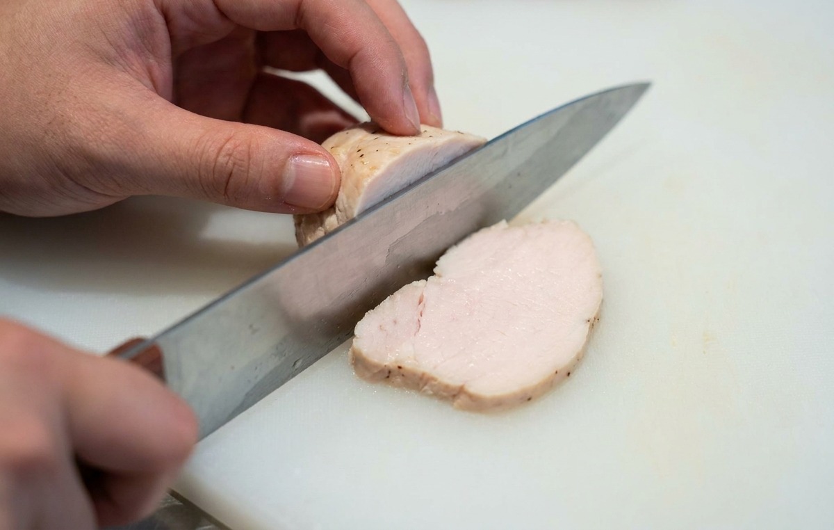 Mani che affettano un pezzo di petto di pollo cotto a bassa temperatura in fette sottili su un tagliere bianco.