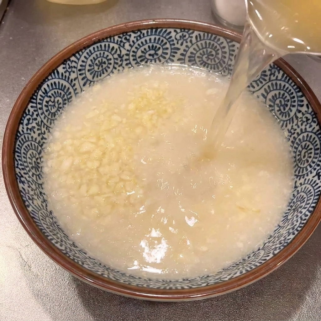 Brodo caldo e limpido versato da una pentola in una ciotola di ceramica contenente aglio grattugiato.