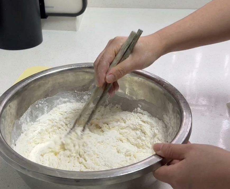 Una persona usa delle bacchette di legno per mescolare farina bianca e acqua in una ciotola di acciaio inossidabile.