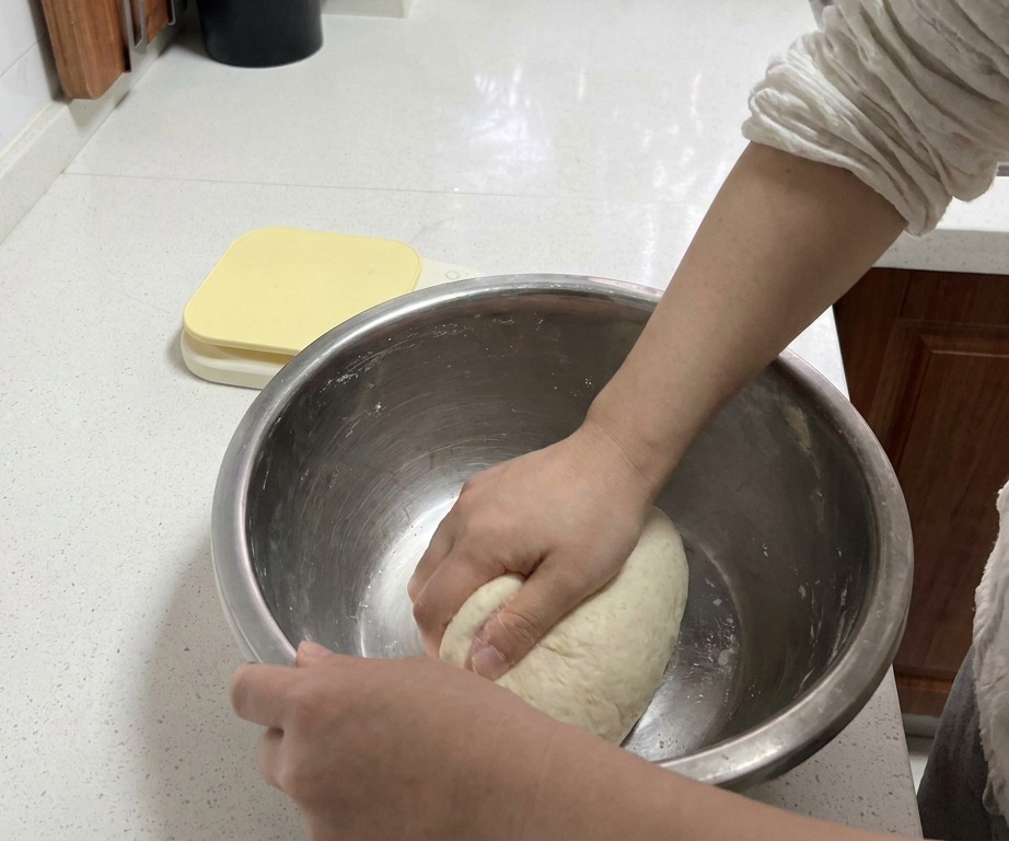 Mani che impastano con decisione una palla di pasta bianca in una ciotola di metallo su un piano di lavoro pulito.