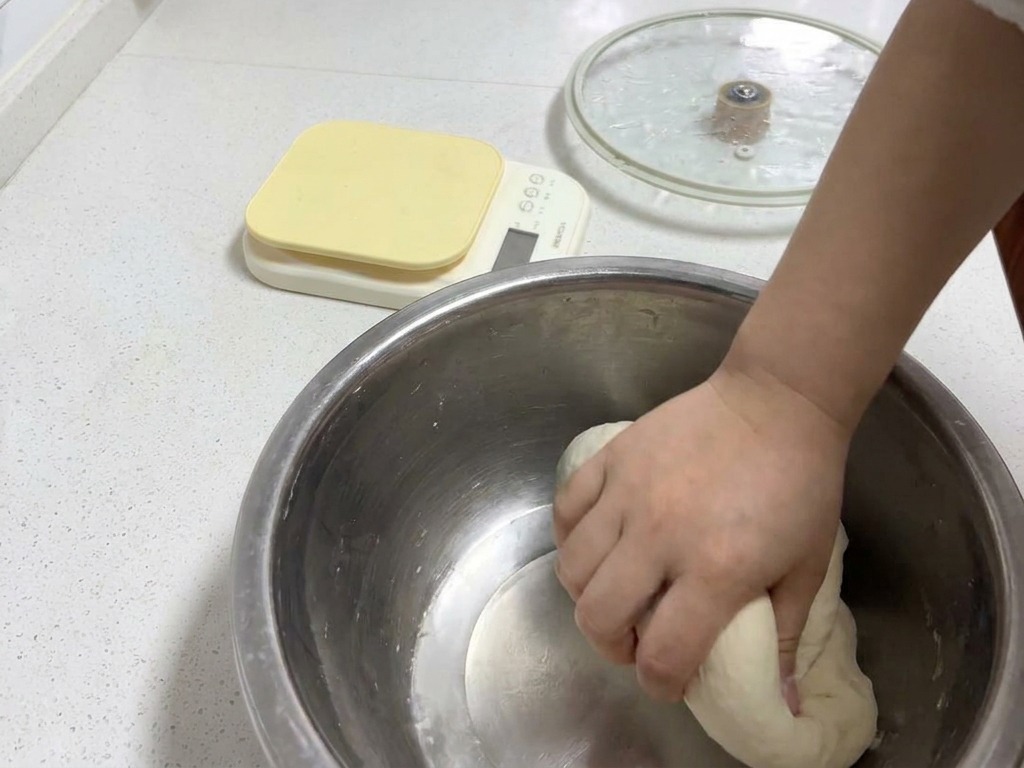 Una mano che impasta una palla di pasta liscia e rotonda in una ciotola di metallo per sviluppare una consistenza elastica.