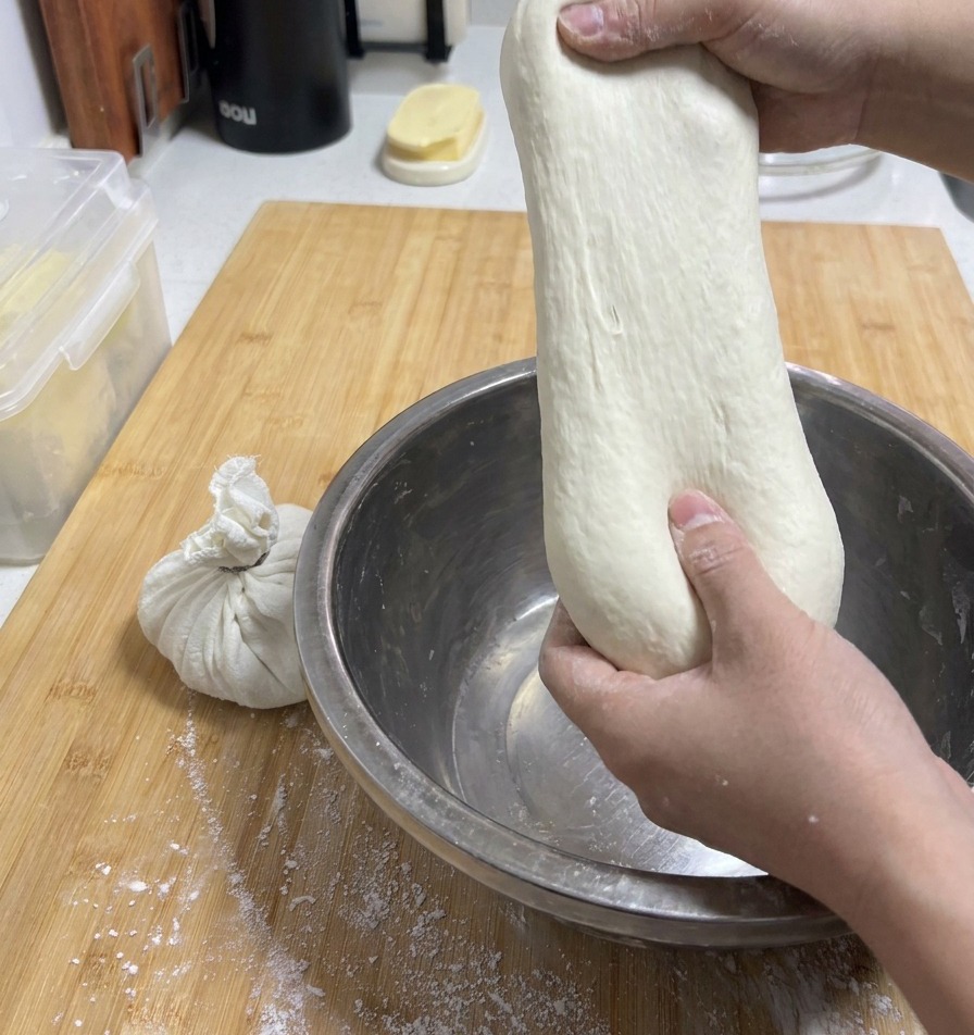 Mani che tirano un pezzo di pasta liscia e chiara verso lalto da una ciotola per mostrarne lincredibile elasticità.