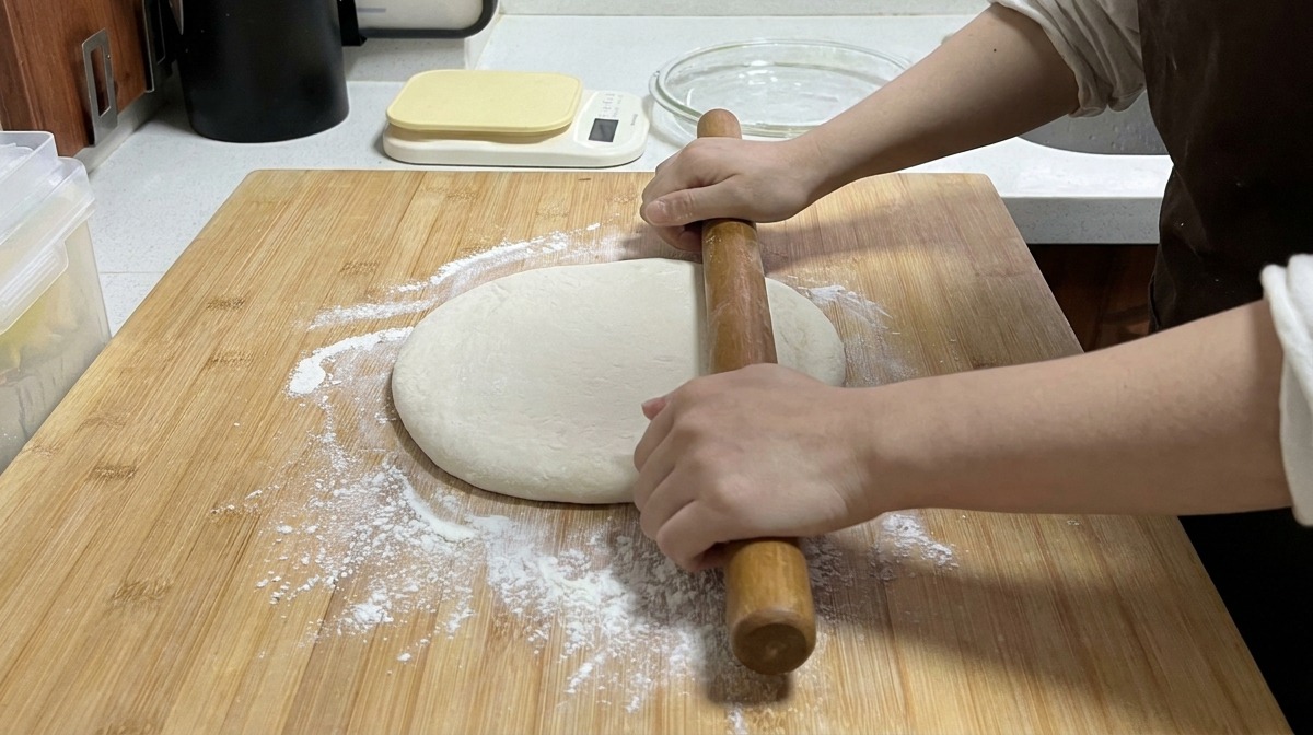 Mani che usano un lungo mattarello di legno per appiattire limpasto su un tagliere di bambù infarinato.