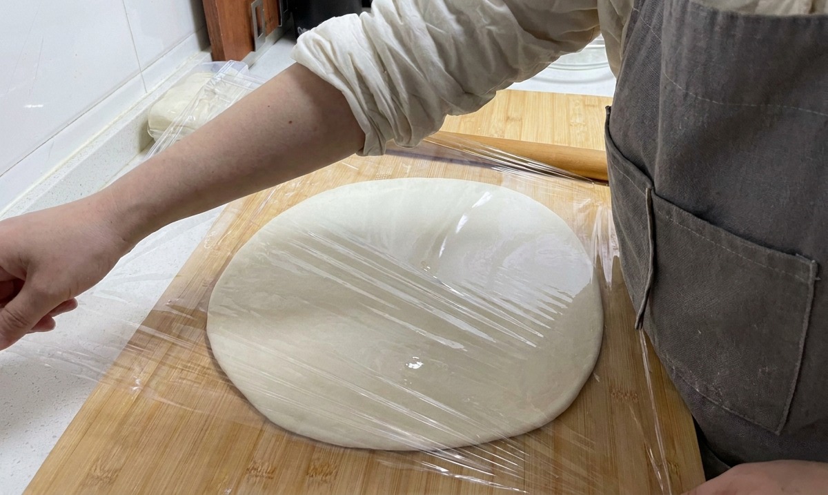 Una mano che stende della pellicola trasparente sopra un grande disco di pasta su un tagliere di legno.