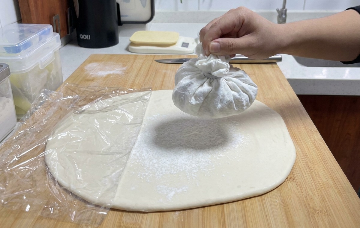 Una mano che scuote un sacchetto di stoffa per rilasciare un velo di farina bianca sopra la sfoglia di pasta.